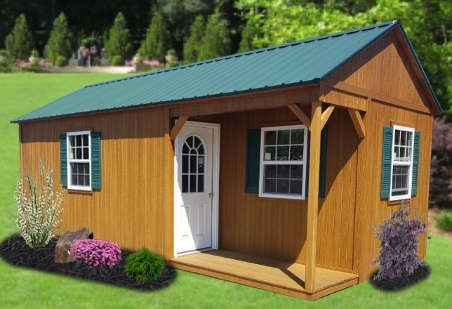 A small wooden shed with a green metal roof and white door sits on grass. It has blue shutters, surrounded by shrubs and colorful flowers, under a sunny sky.