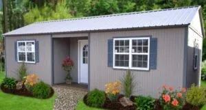 Small gray cottage with white trim and blue shutters. Front door is flanked by flower arrangements and shrubs. Gravel path leads to entrance, surrounded by trees.
