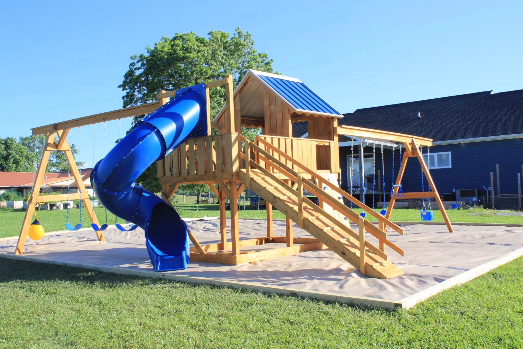 Wooden playground set with blue slide and swings on lawn. Includes a small playhouse with stairs. Sunny day with clear sky and trees nearby.