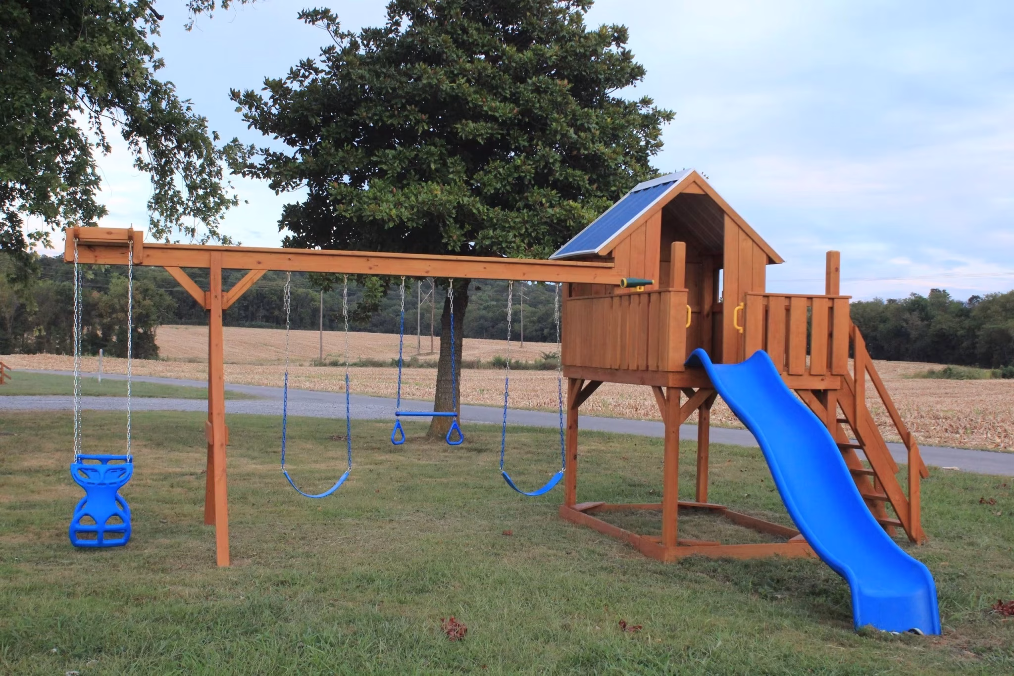 Wooden playset with a slide, swings, and a clubhouse stands on a grassy field under a tree. The vibrant blue slide adds a playful touch.
