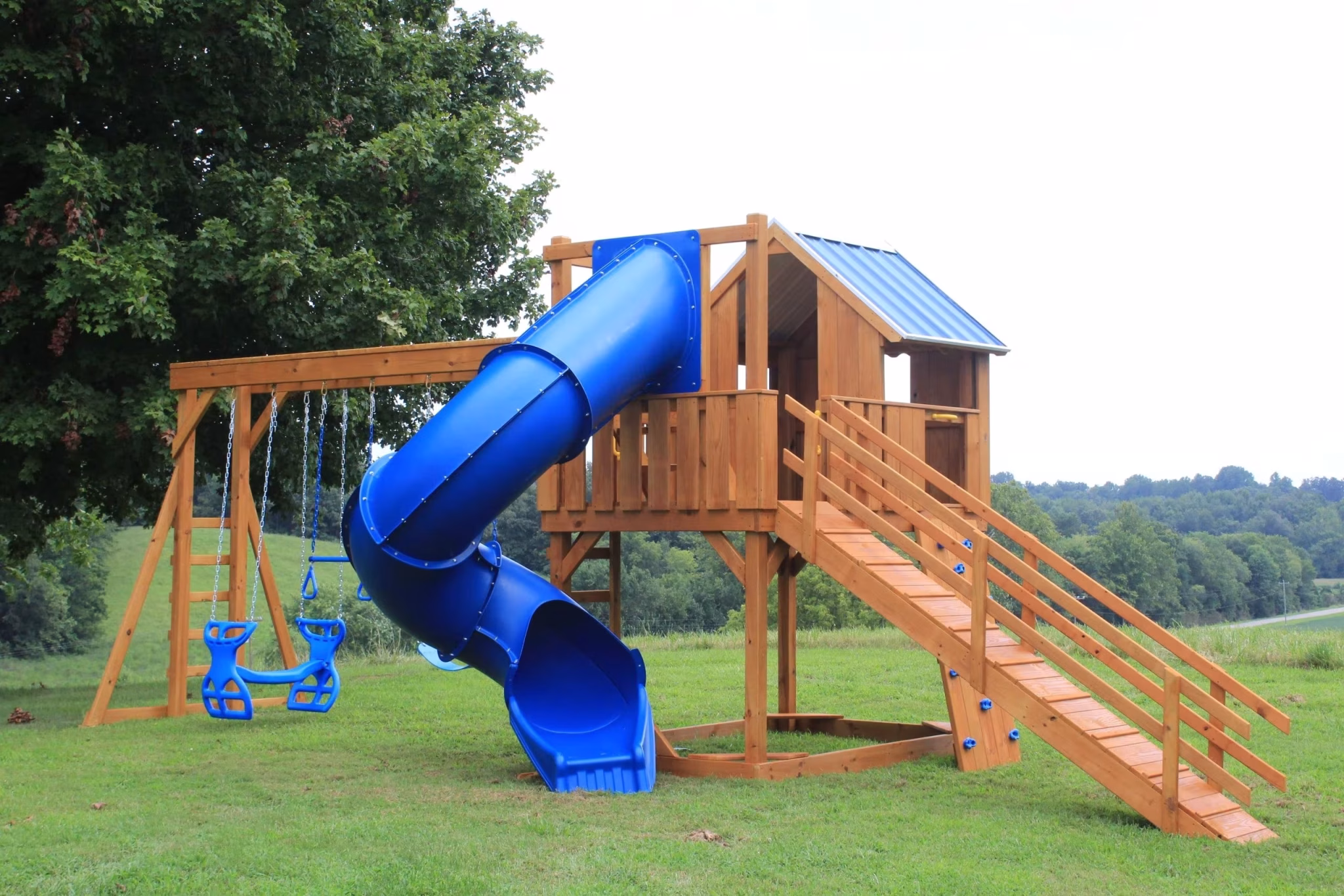 Wooden playset in a grassy area with a blue spiral slide, two blue swings, and a small covered platform. The backdrop is an open field and trees.