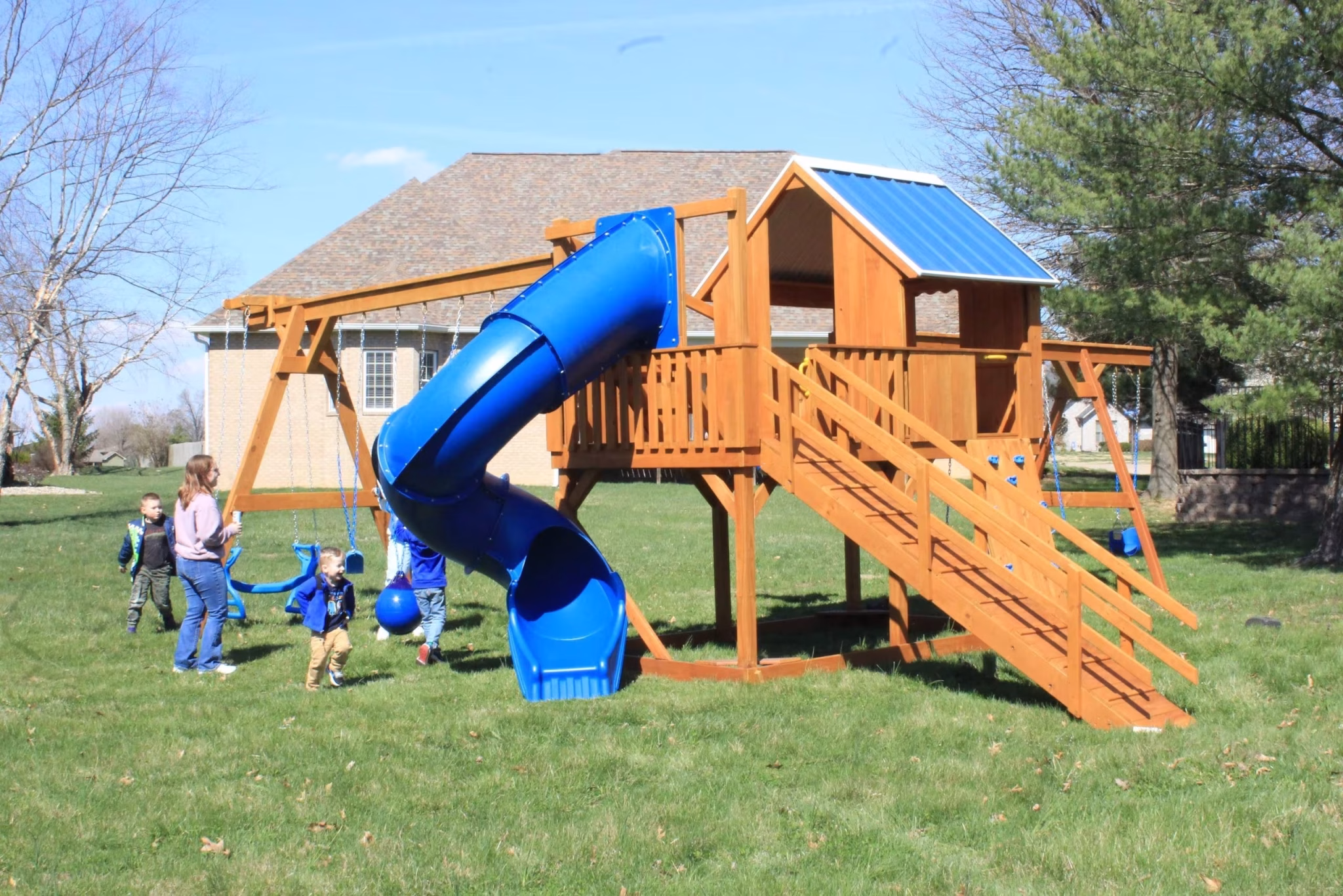 Wooden playset in a grassy area with a blue spiral slide, two blue swings, and a small covered platform. The backdrop is an open field and trees.