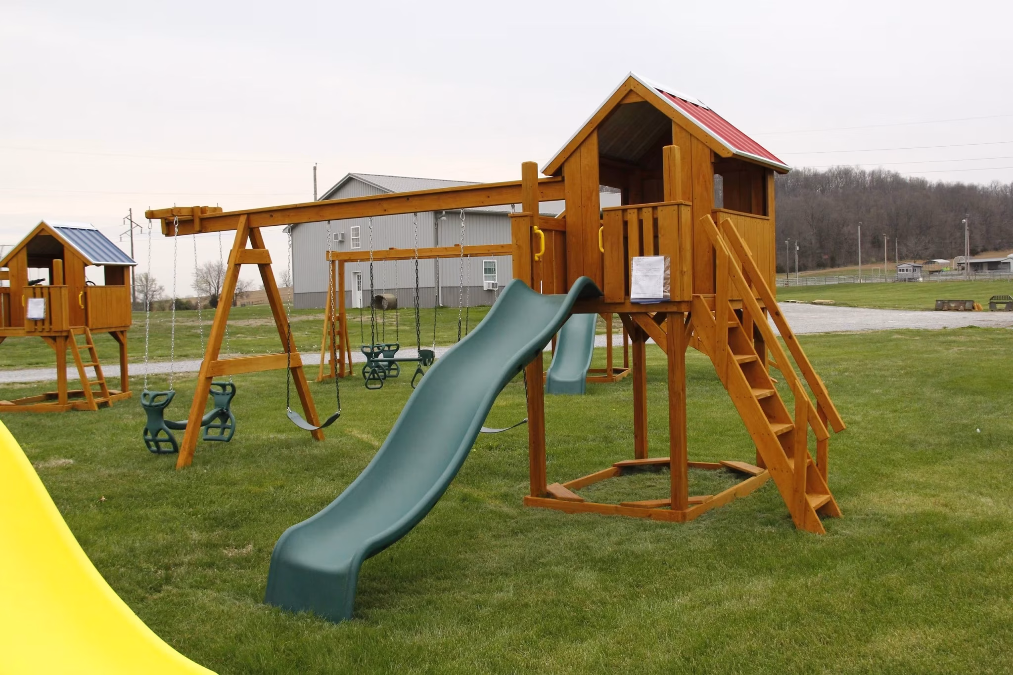 Wooden playground set with a slide, swings, and two raised playhouses on a grassy field. Overcast sky and a building in the background.