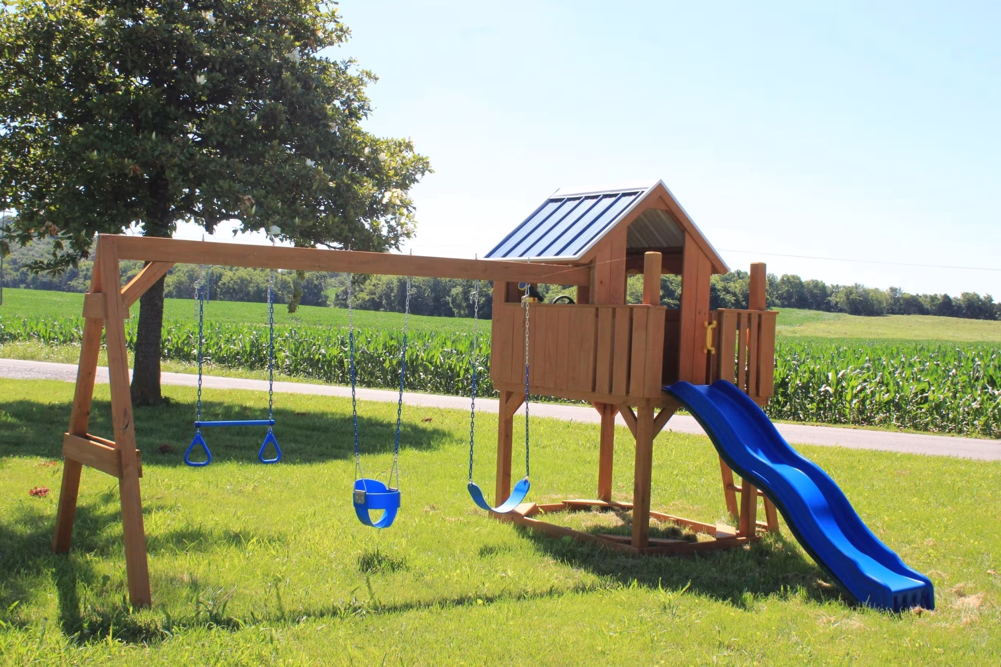 Wooden playset with a slide, swings, and a clubhouse stands on a grassy field under a tree. The vibrant blue slide adds a playful touch.