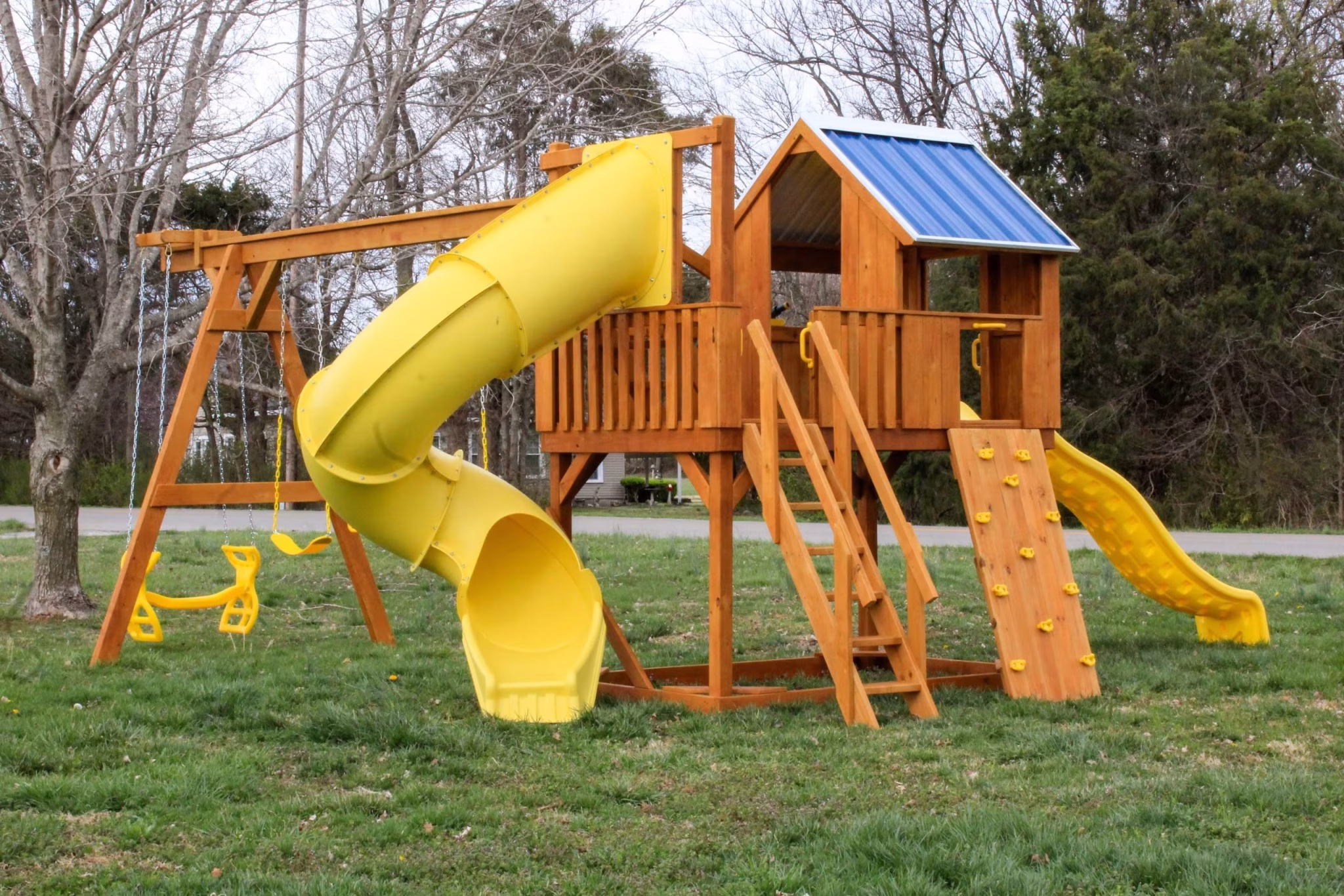 Wooden playground set with blue-roofed playhouse, yellow tube slide, wave slide, swings, and climbing wall on grass, surrounded by bare trees.