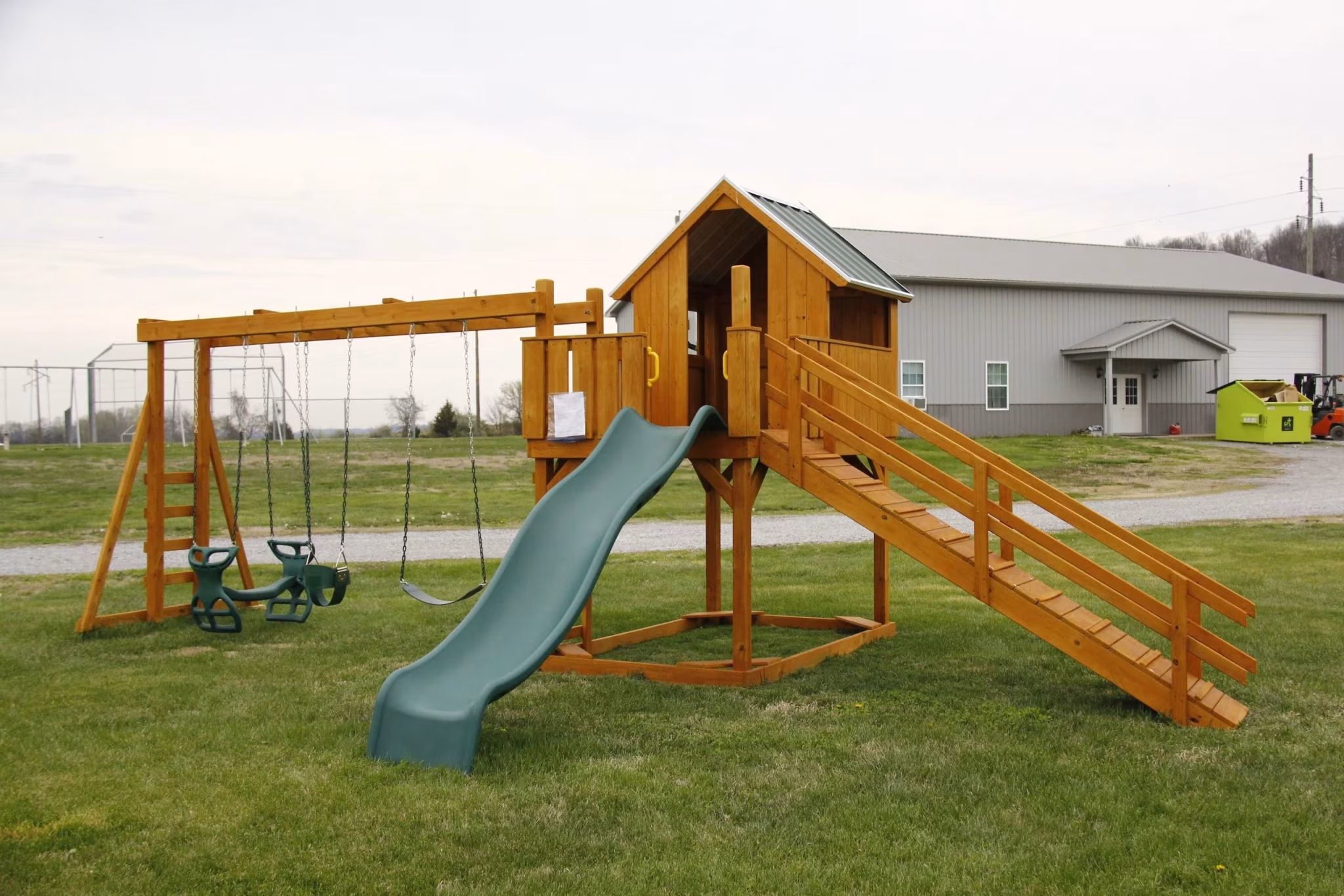 Wooden playground set with a slide, swings, and two raised playhouses on a grassy field. Overcast sky and a building in the background.
