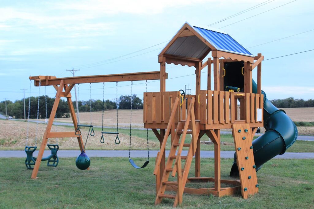 Wooden playground set with a slide, swings, and two raised playhouses on a grassy field. Overcast sky and a building in the background.