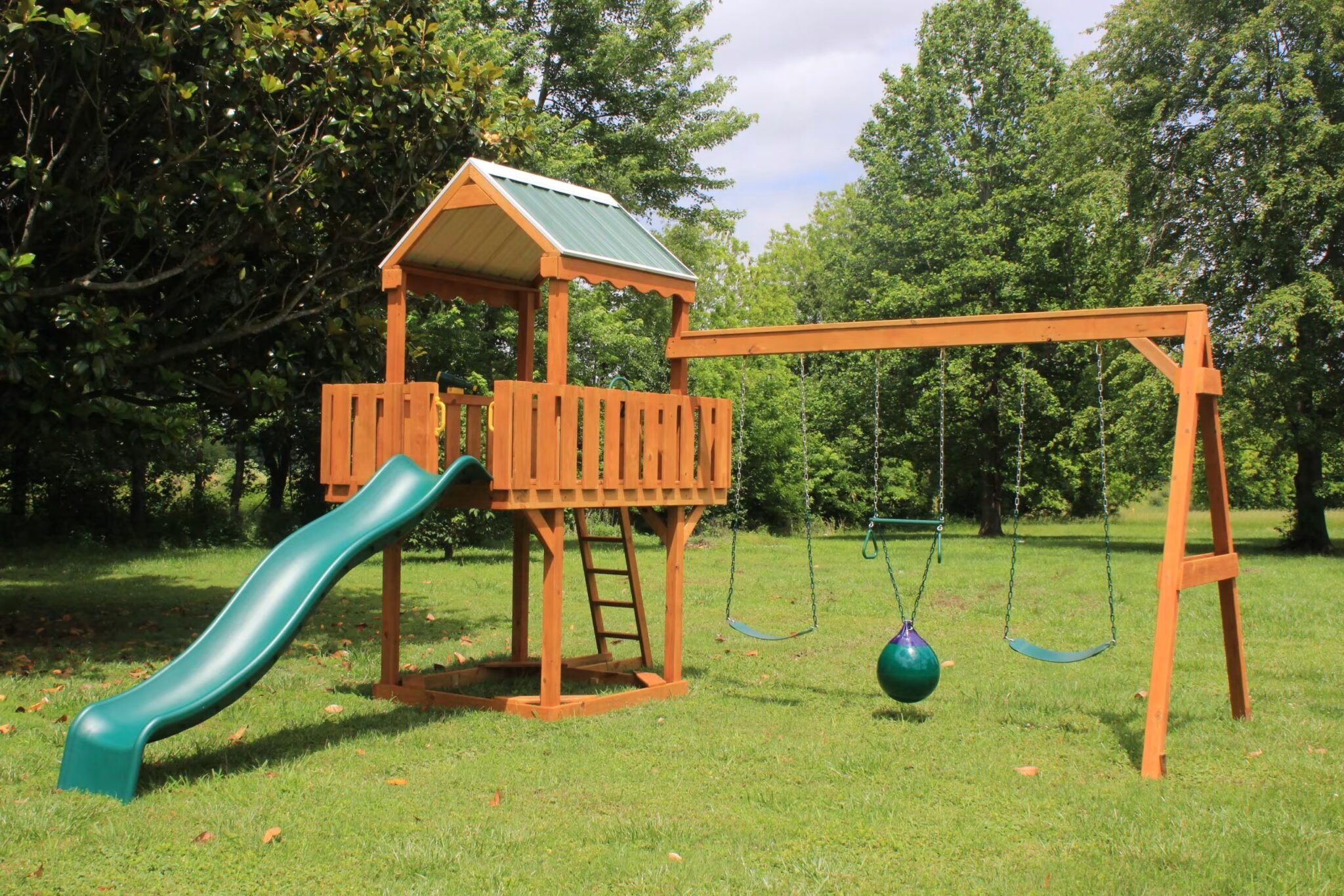 Wooden playground set with a slide, swings, and two raised playhouses on a grassy field. Overcast sky and a building in the background.